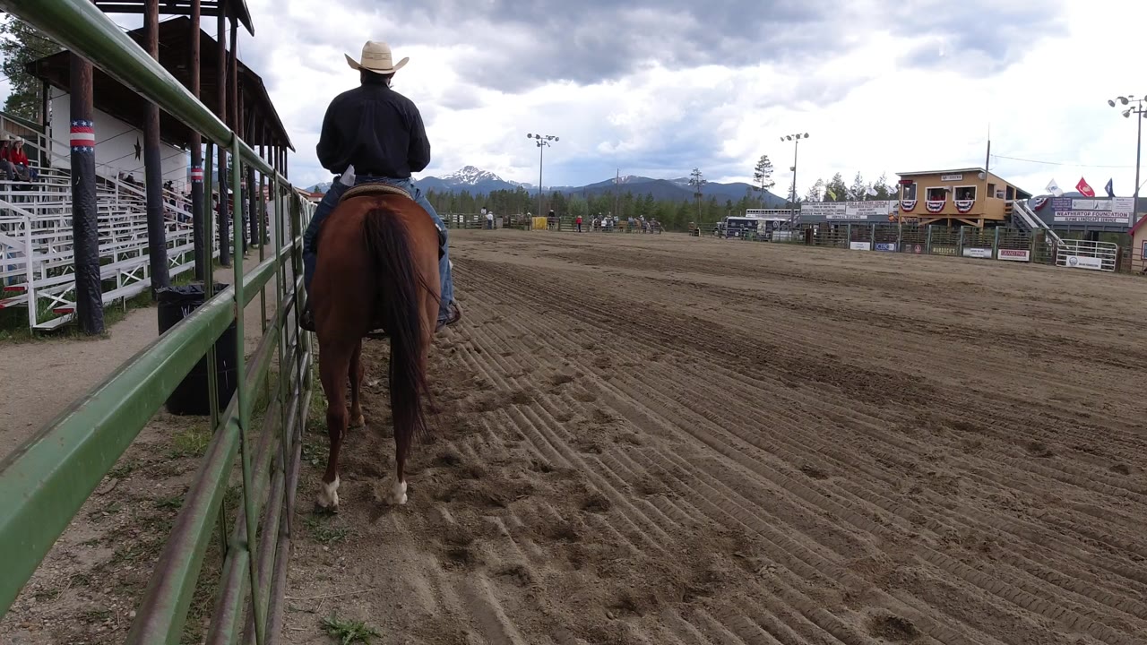 A rider in a light cowboy hat sits on a brown horse near green arena rails in Winter Park, Colorado, with dirt track, bleachers, and cloudy mountains beyond.