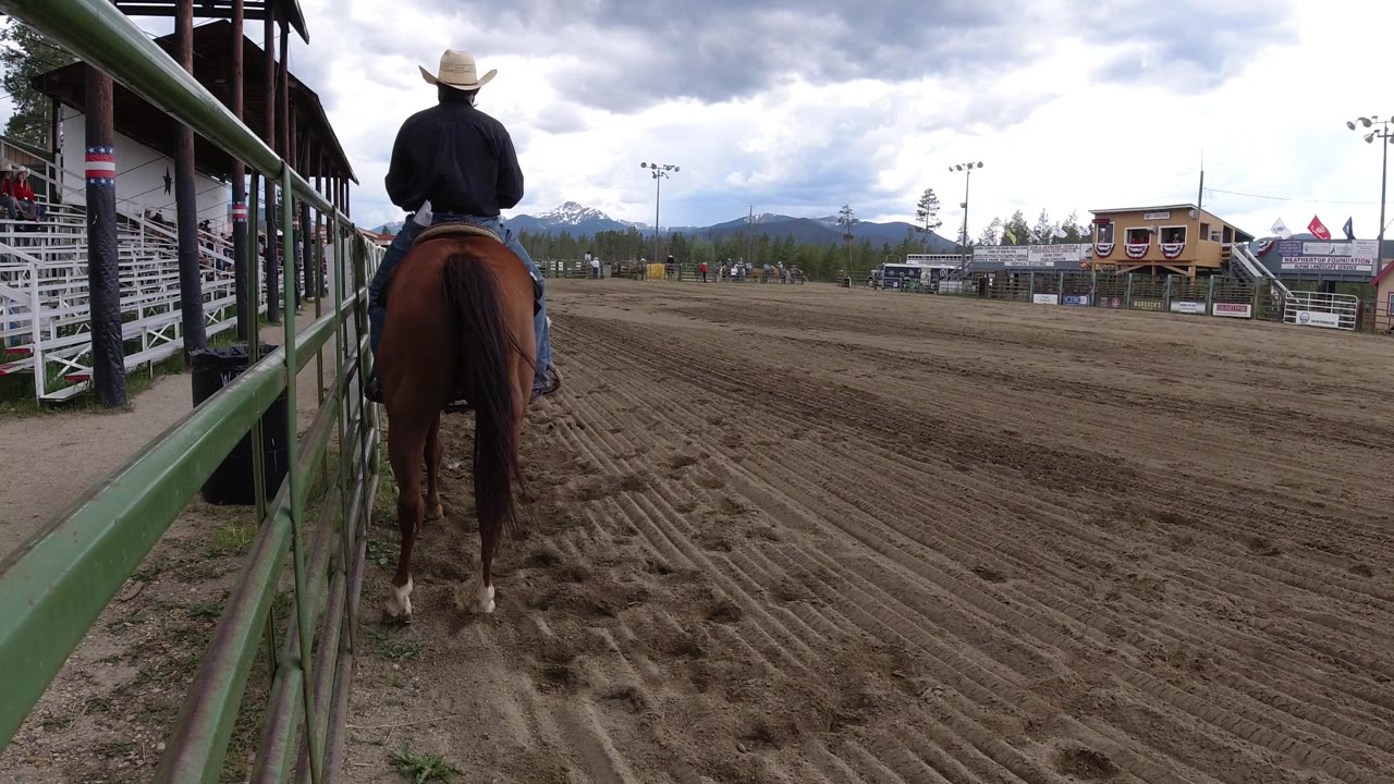 The horse and rider remain near the left rail in Winter Park, Colorado, looking across the empty dirt arena toward bleachers, buildings, trees, and mountains.