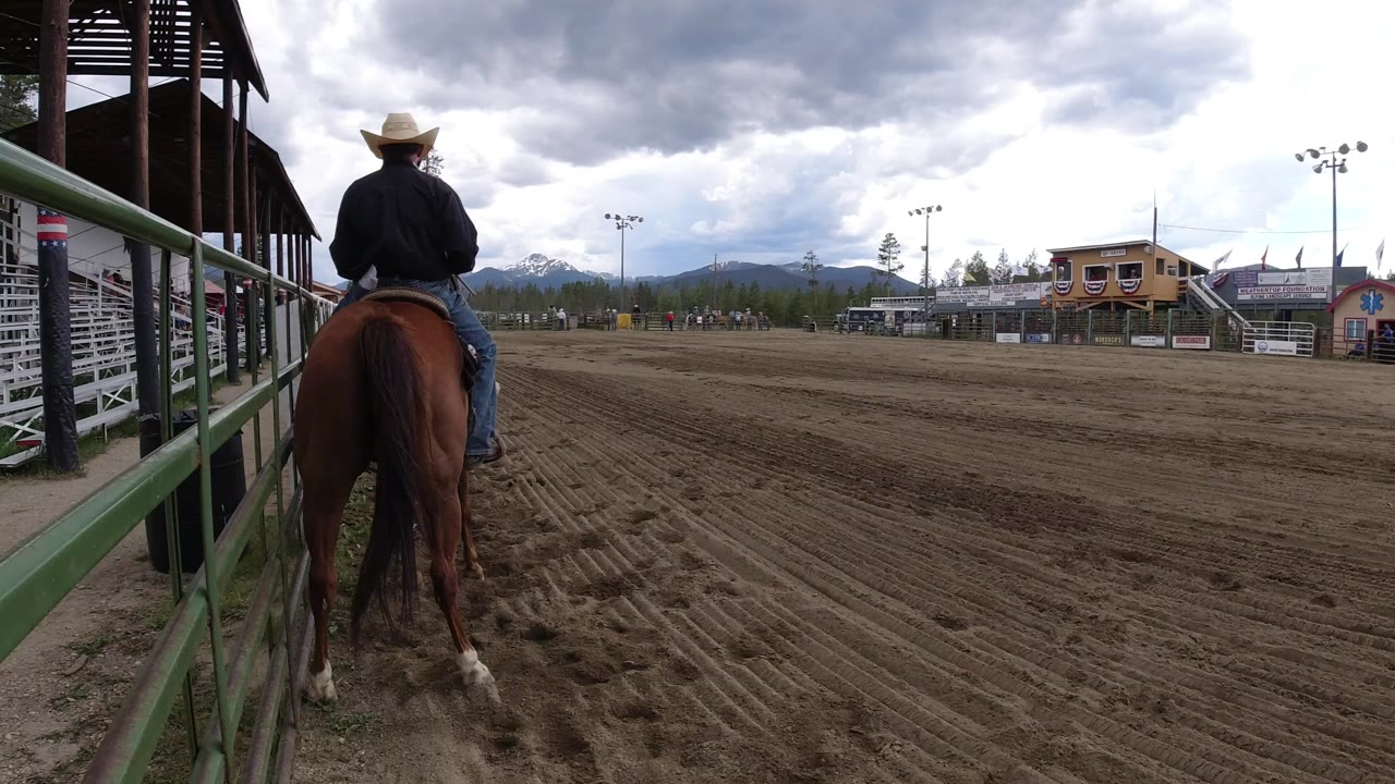 In Winter Park, Colorado, a rider in a cowboy hat sits on a brown horse at the edge of a dirt arena, with bleachers and distant mountains under cloudy sky.