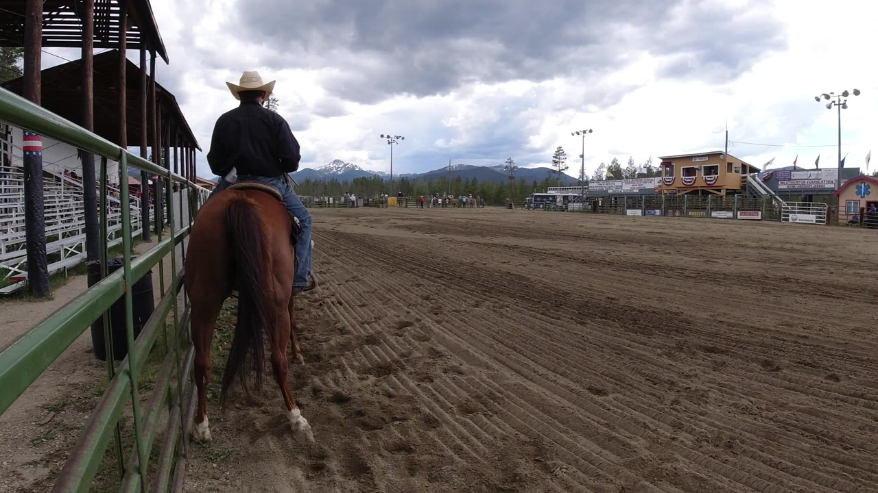 The rider and brown horse remain near the green rail in a Winter Park dirt arena, facing toward bleachers, arena buildings, and mountains in the distance.