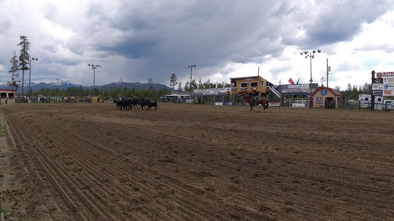 Winter Park, Colorado dirt arena seen from a wide ground-level view, with horses and riders in the middle distance, event buildings behind them, mountains on the horizon, and people visible.