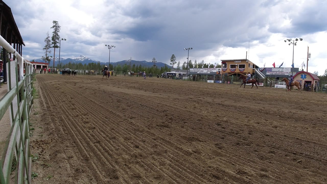Riders on horses pass near arena buildings with mountains behind them.