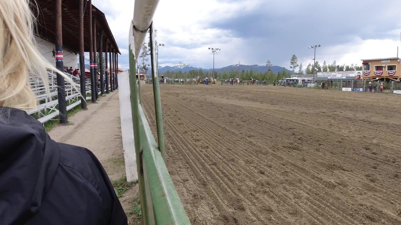 Dirt horse track beside a green rail with bleachers and mountains in the distance.