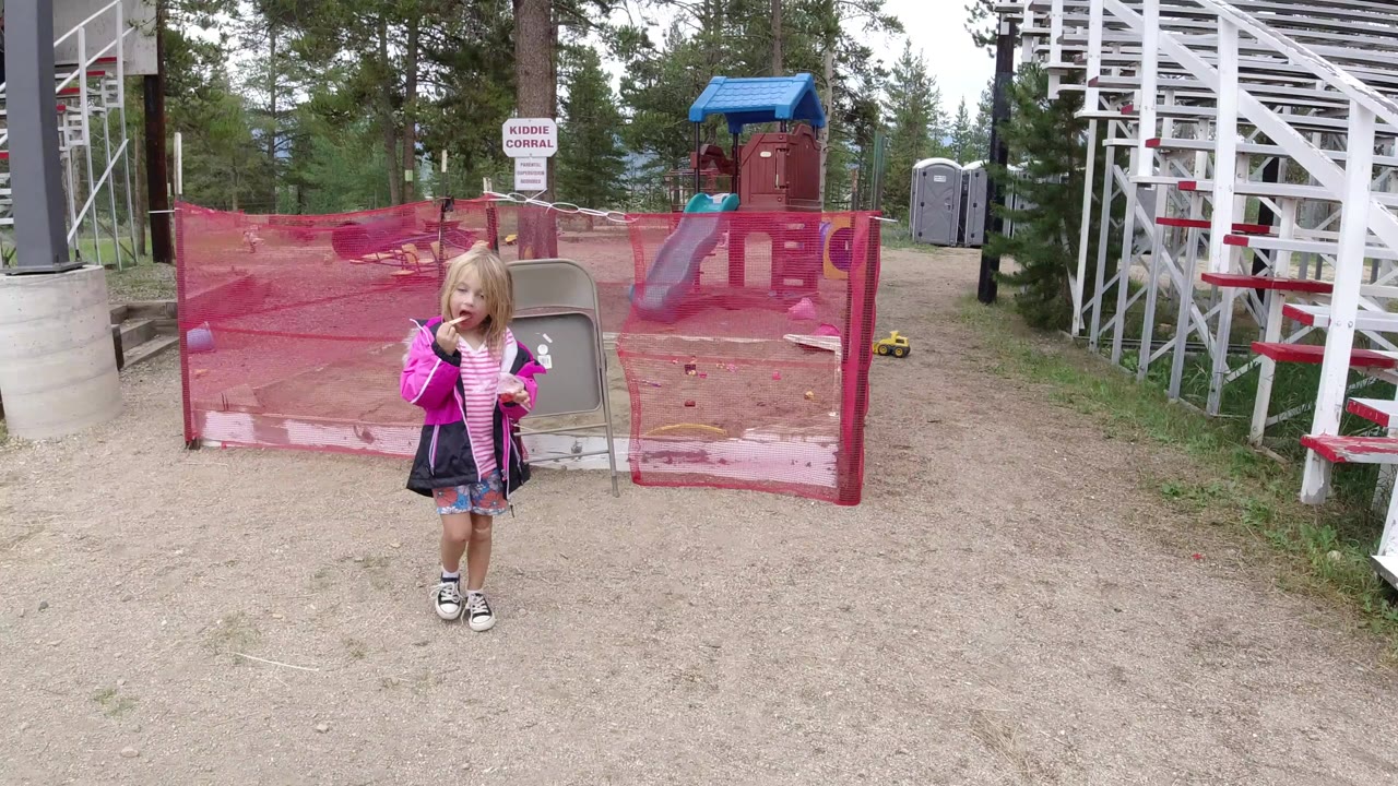 Young blonde girl in a bright pink jacket stands on gravel near red mesh corral fencing, a small play structure, white bleachers, and trees in Winter Park, Colorado.