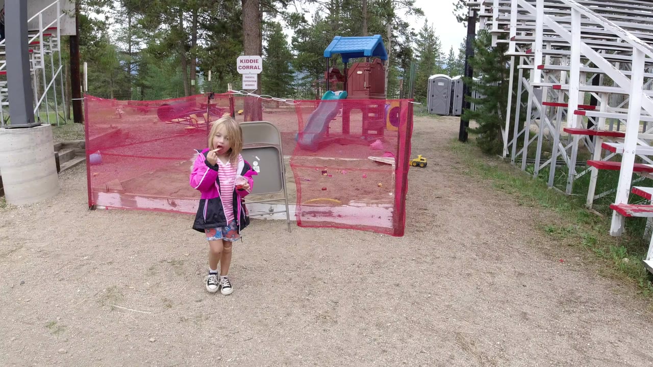 The girl remains centered in front of the red mesh fence, with white grandstand seating to the right and a small sign behind the fenced area in Winter Park, Colorado.