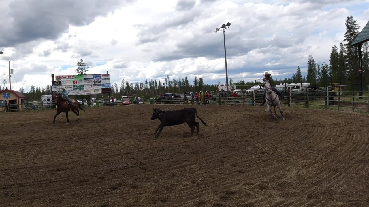 In Winter Park, Colorado, two horseback riders flank a black cow in a dirt rodeo arena beneath a cloudy sky, with sponsor boards and fencing in the background.