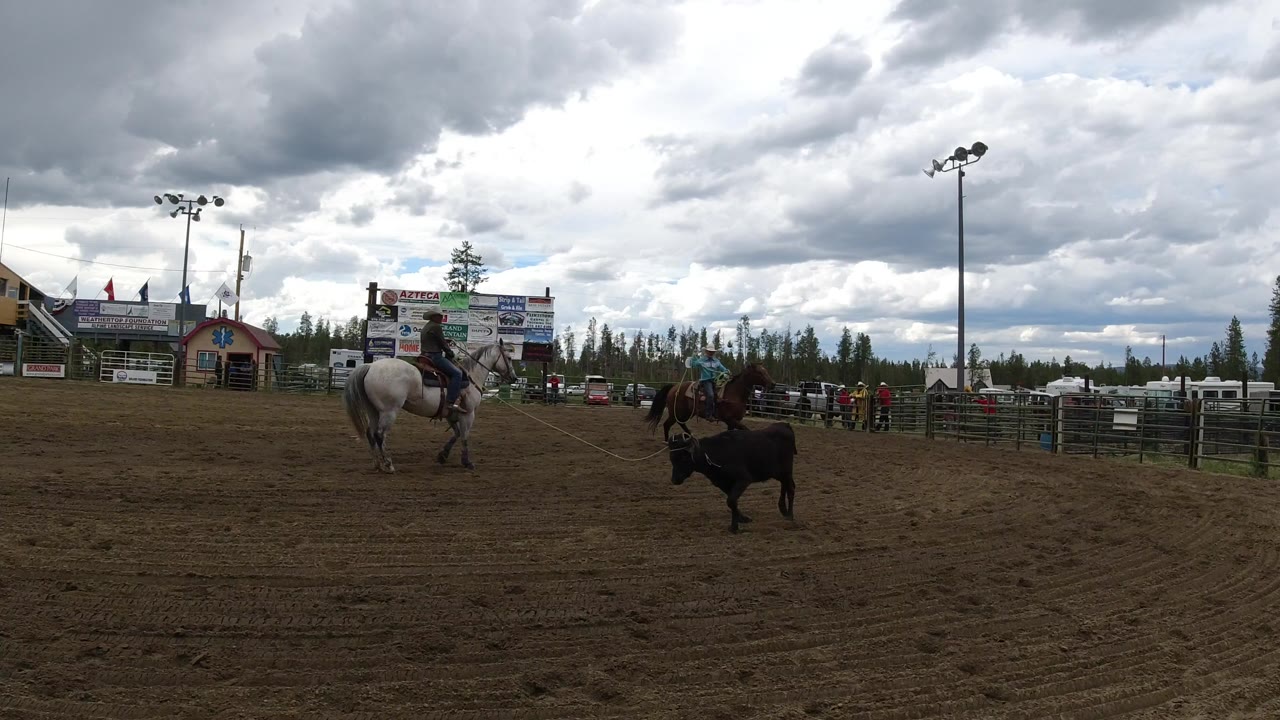 Two distant mounted riders flank a black calf in a Winter Park, Colorado dirt arena, with bleachers, sponsor boards, pine trees, and no close-up faces visible.