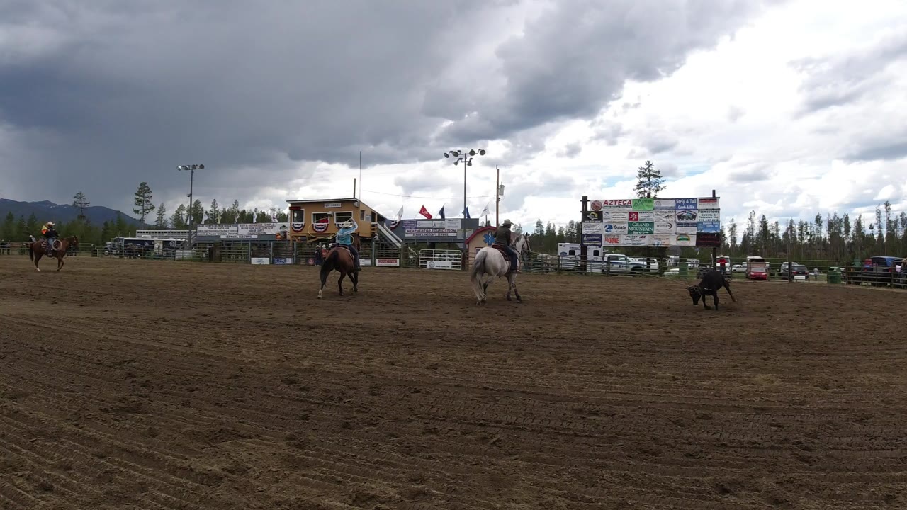 Wide rodeo arena with mounted riders, a black bull, grandstands, and storm clouds.