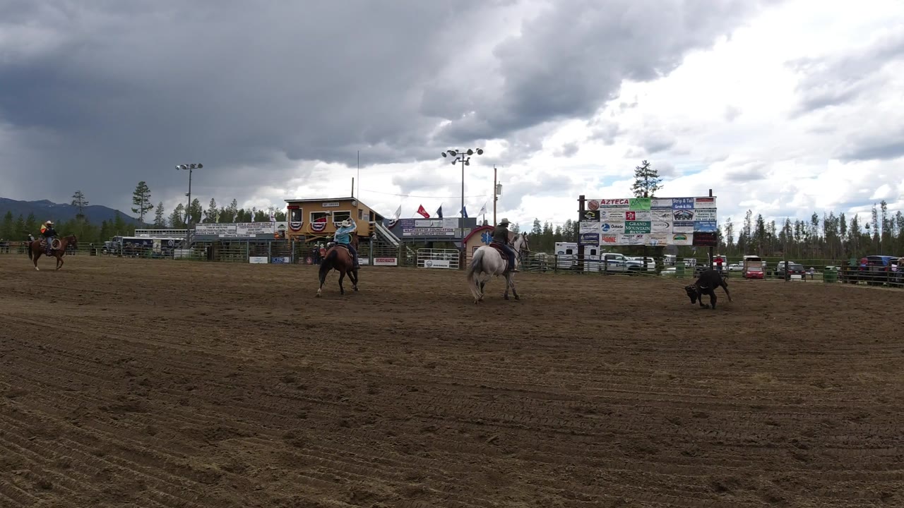 In the next moment, the black bull moves through the midground of the Winter Park, Colorado arena while riders on horses hold distance near the bleachers, sponsor boards, pine trees, and cloudy mountain horizon.