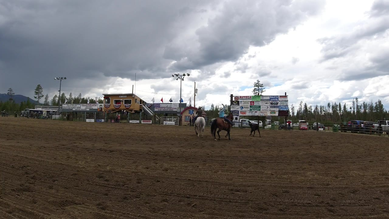 Wide view of a dirt arena in Winter Park, Colorado, with two horses and distant riders near fencing, sponsor boards, parked vehicles, trees, mountains, and heavy clouds.