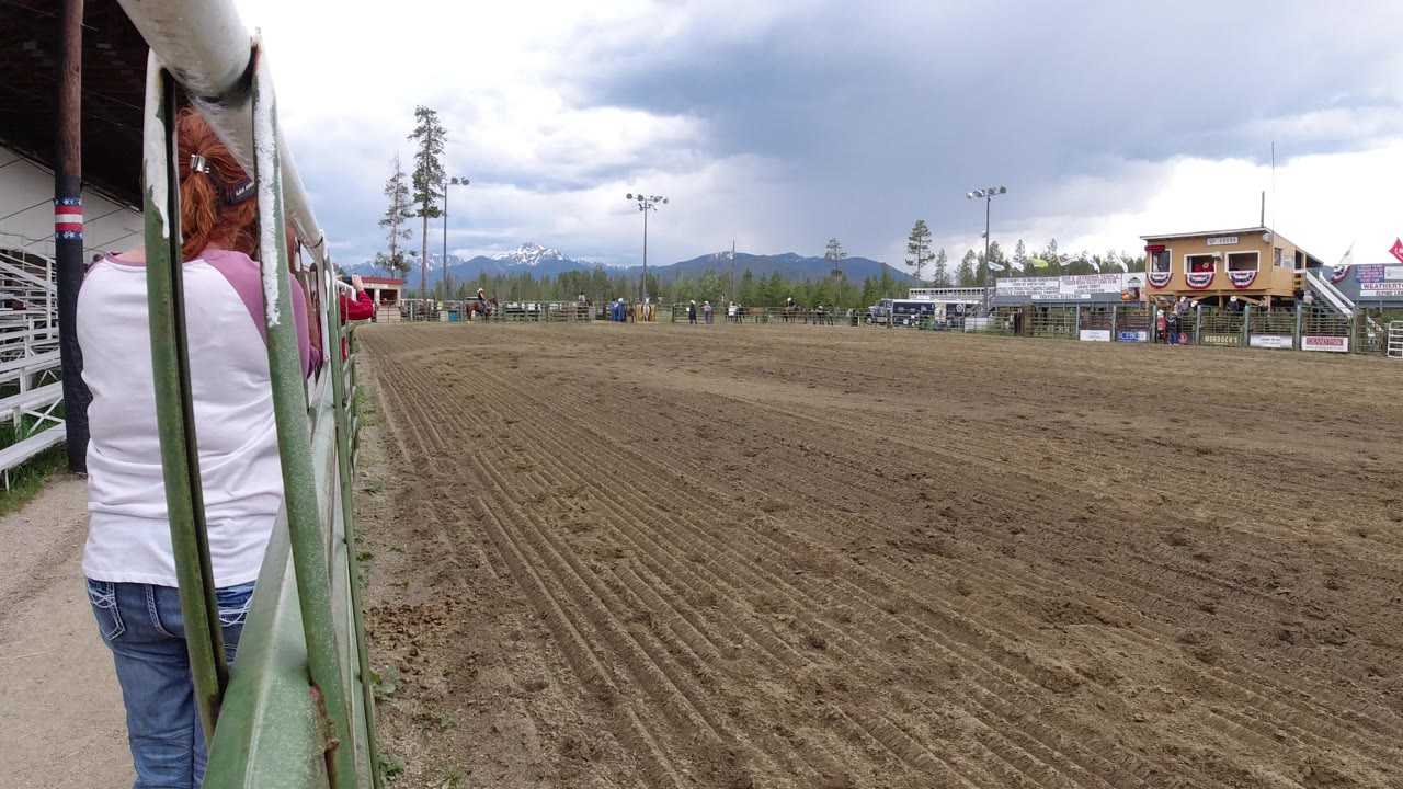 Woman standing by green railings beside a dirt arena under cloudy mountains.