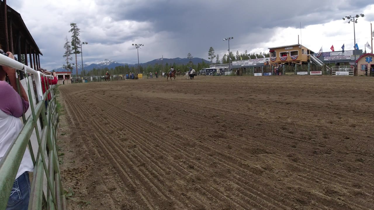 Winter Park, Colorado rodeo view with riders and a calf moving across the far side of the dirt arena, a partial spectator by the rail, small buildings, flags, trees, and dark clouds.