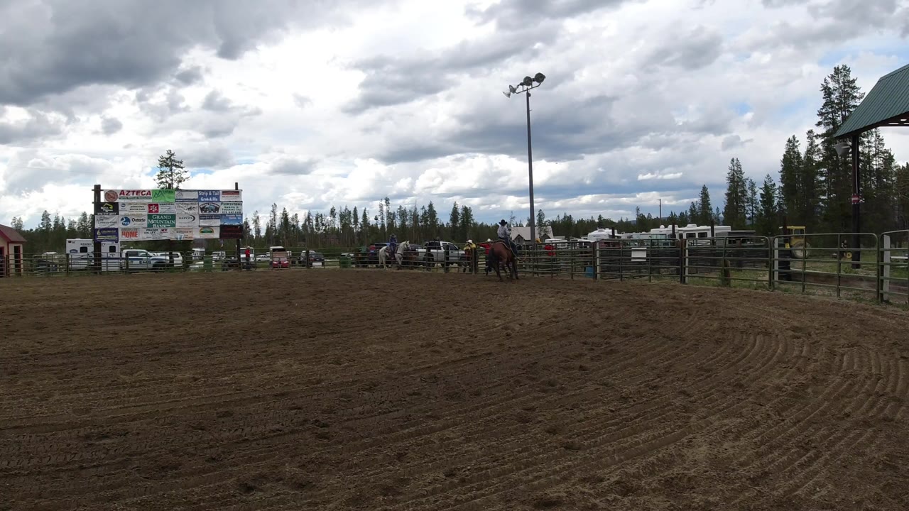 Wide view of a dirt arena in Winter Park, Colorado with rail fencing, distant horses and riders, sponsor boards, evergreen trees, arena lights, and a heavy cloudy sky.