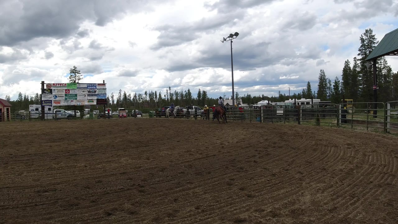 Winter Park, Colorado arena view with raked dirt foreground, low fencing, small distant riders, parked vehicles, sponsor signage, evergreens, and a tall light pole under gray clouds.