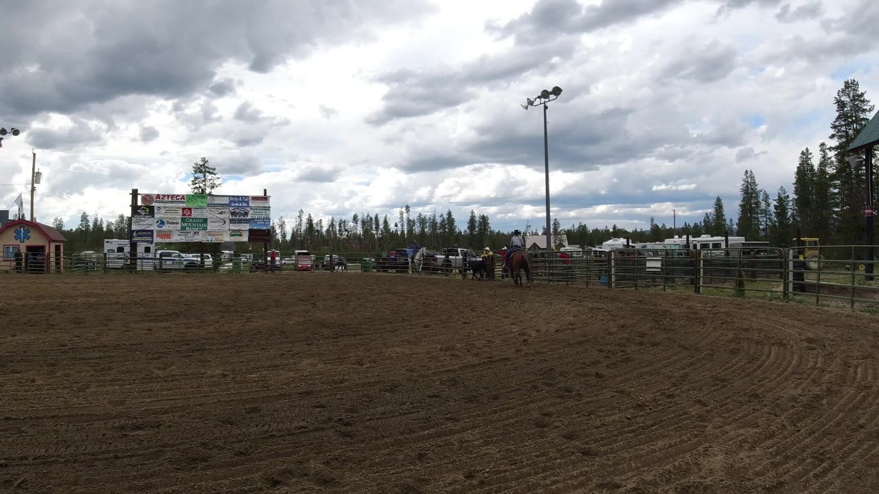 Outdoor arena with sponsor boards, horses near fencing, vehicles, trees, and clouds.