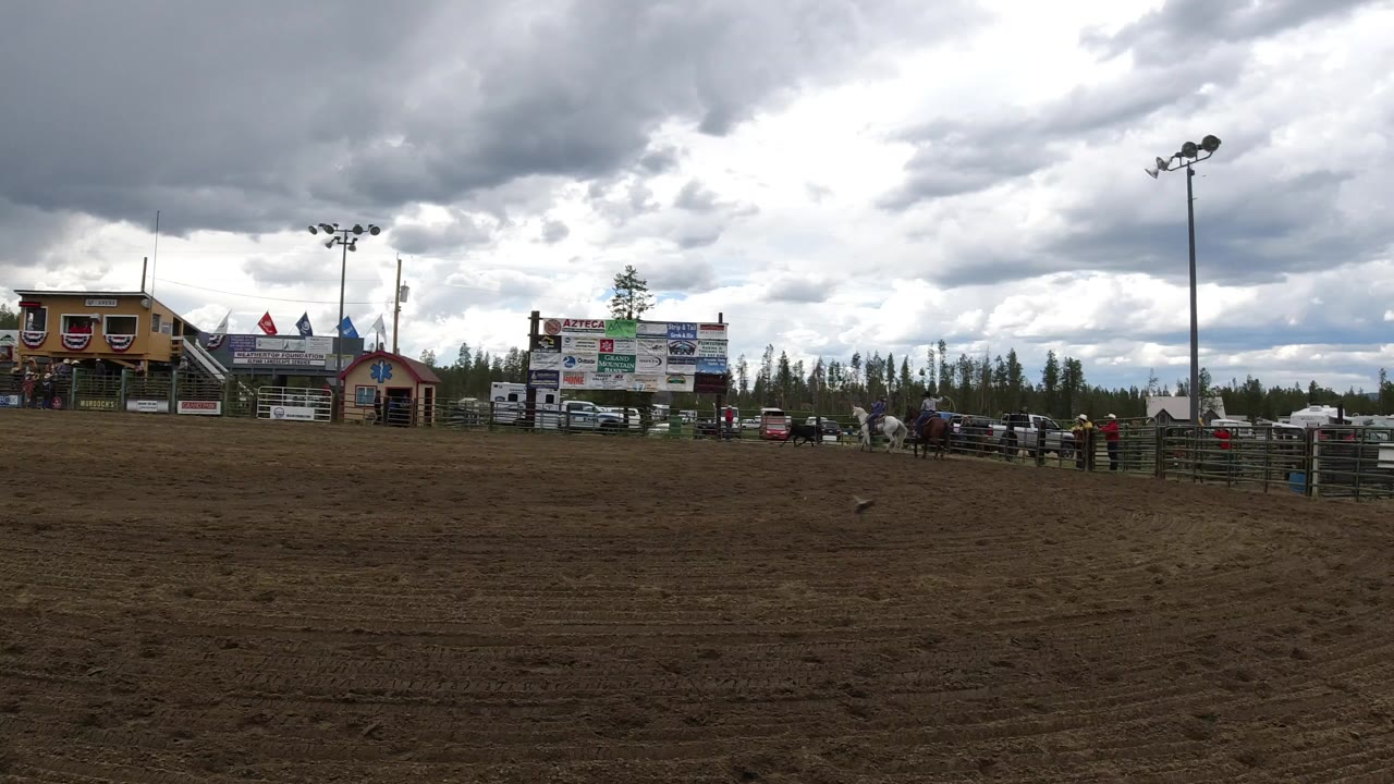 Wide dirt arena with buildings, signs, fences, trees, and storm clouds.