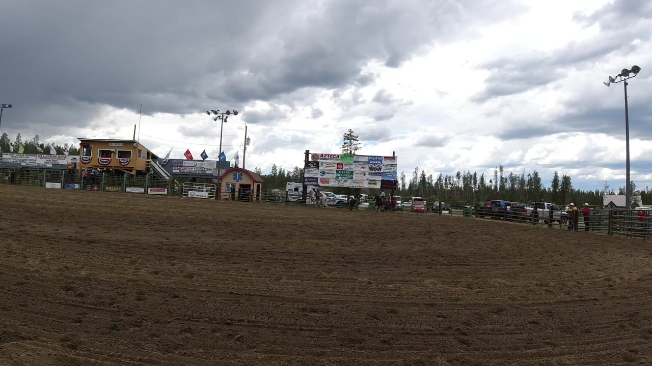 Opening wide view of a muddy arena in Winter Park, Colorado, with small distant riders near the fence, event buildings, signs, flags, parked vehicles, pine trees, and heavy clouds.