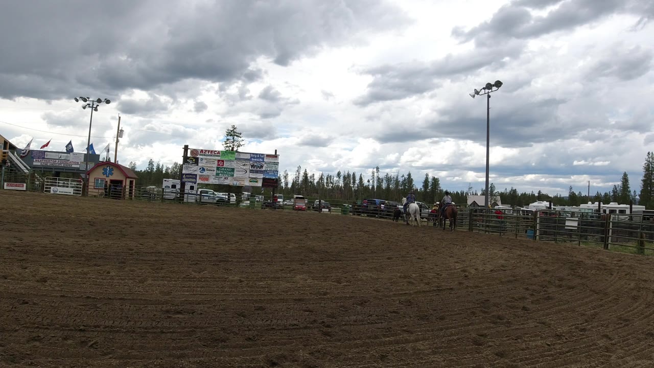 Winter Park, Colorado arena frame with horses clustered near the right fence, low buildings and sponsor-style signboards in the midground, and dark clouds overhead.