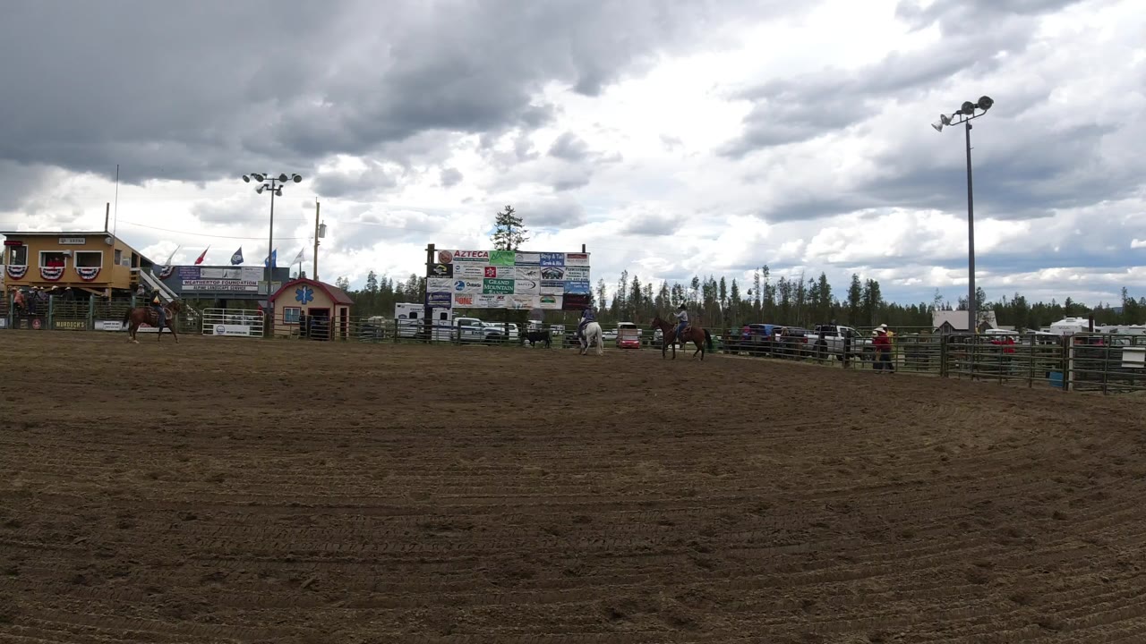 Wide view of a Winter Park, Colorado fairground with a dirt arena in front, horses and riders near the fence, buildings and sponsor boards behind them, and heavy gray clouds overhead.