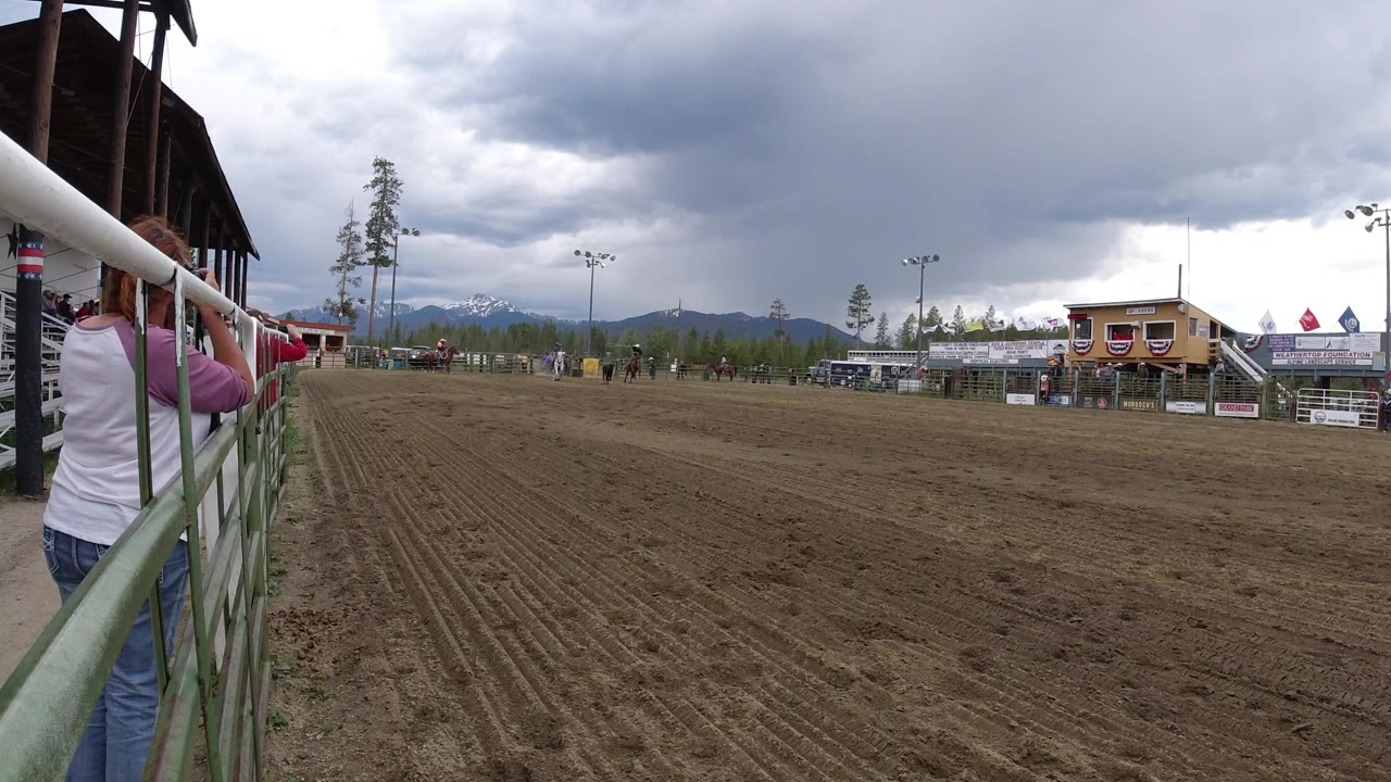 Wide dirt riding arena with rail fence, distant riders, buildings, trees, mountains, and storm clouds.