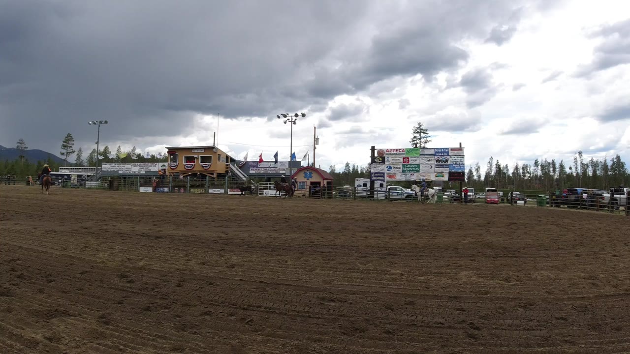 Opening frame in Winter Park, Colorado shows a broad sandy arena with distant horses and riders, low buildings, sponsor-style signs, pine trees, mountains, and dark overcast clouds.