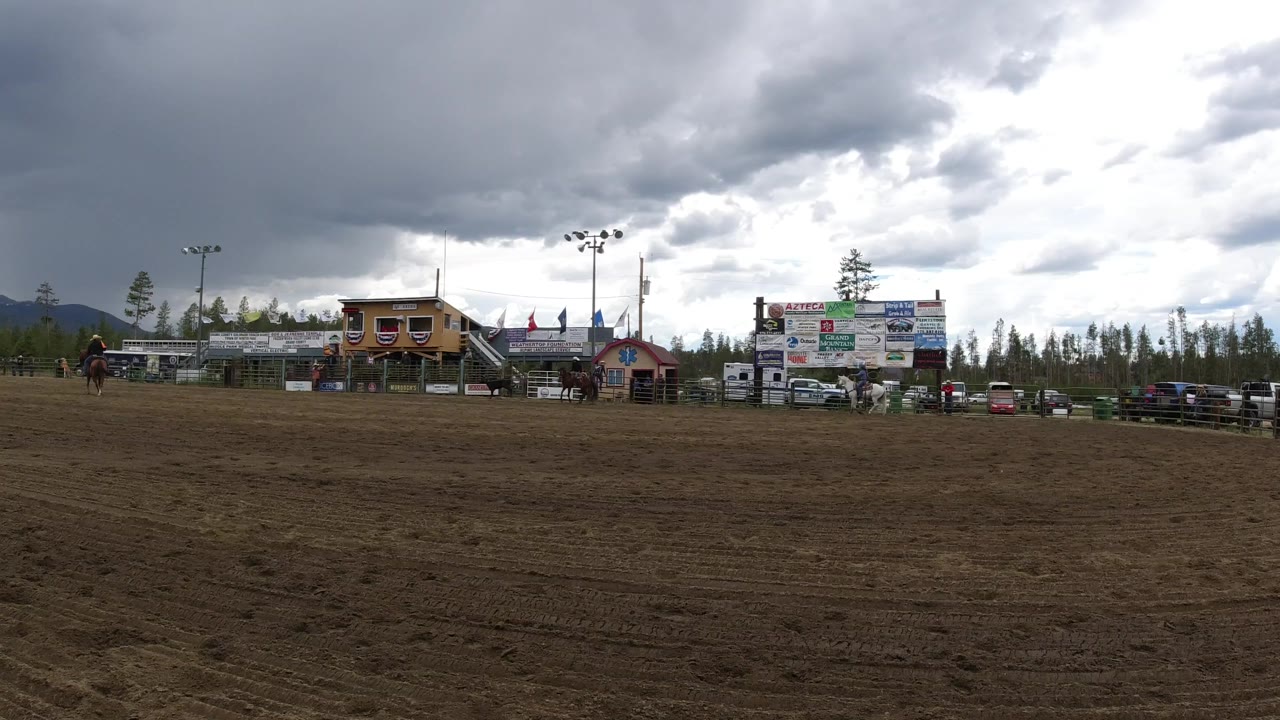 At 0:01, the Winter Park arena remains in a wide view, with a horse and rider left of center, buildings and sign boards across the midground, and mountains under heavy clouds.