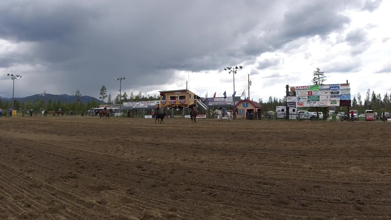 Wide Winter Park, Colorado rodeo arena view with a dirt foreground, two distant horseback riders and a bull near the middle, sponsor boards and trees behind them, and no recognizable faces.