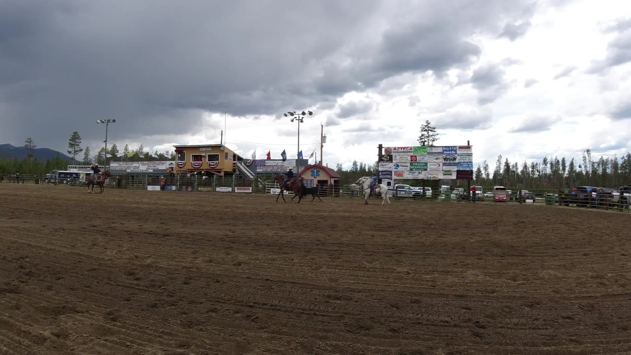 Wide dirt arena with riders, a bull, event buildings, signs, trees, mountains, and storm clouds.