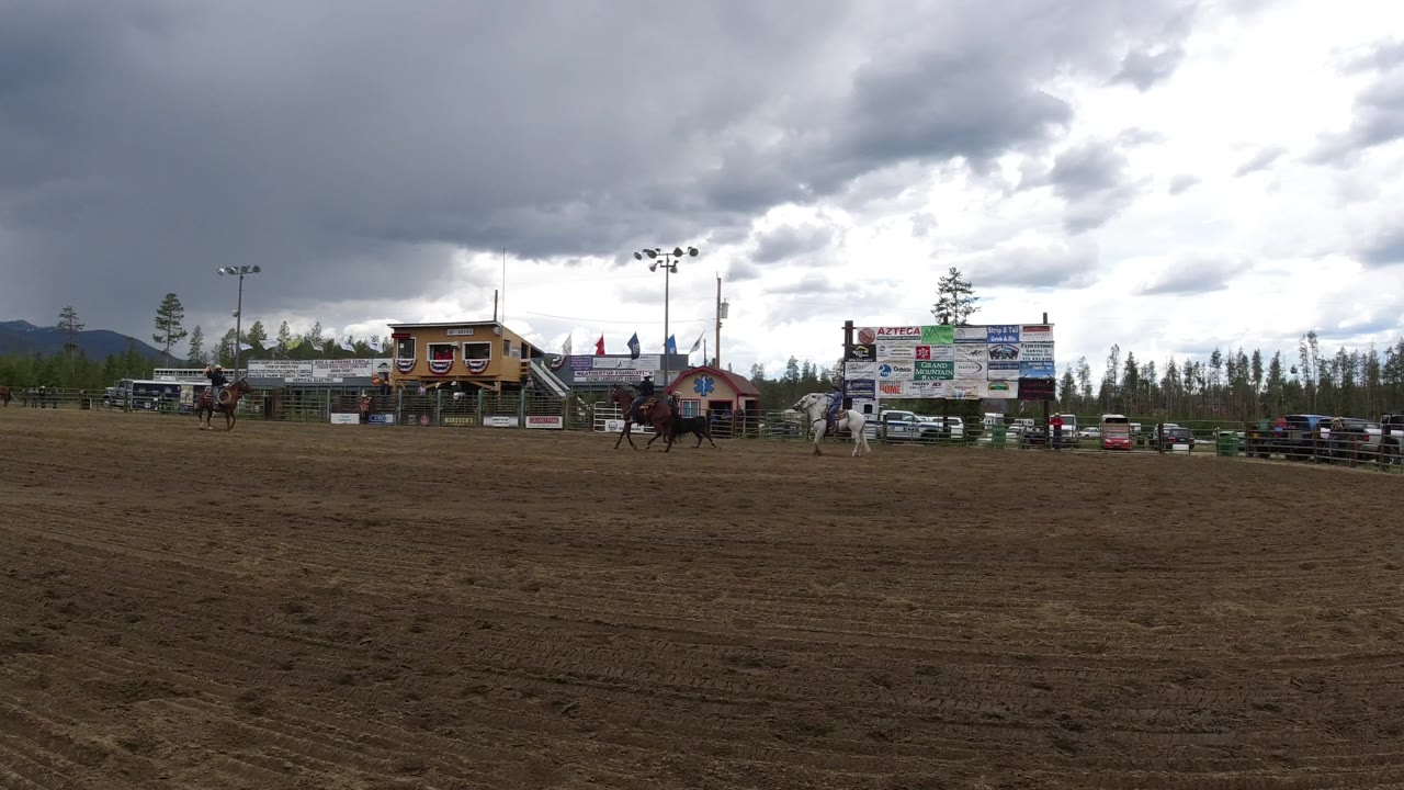 Winter Park, Colorado field with riders spread across the midground, a dark bull near the handling action, and a large sign board beside parked vehicles under broken cloud cover.