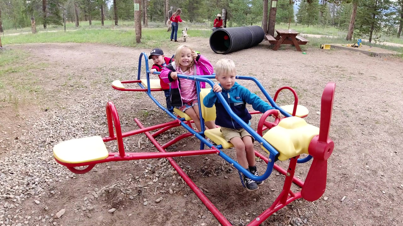 Three children sit on a red, blue, and yellow playground airplane ride in Winter Park, Colorado, with trees, adults, a black tunnel, and picnic tables behind them.