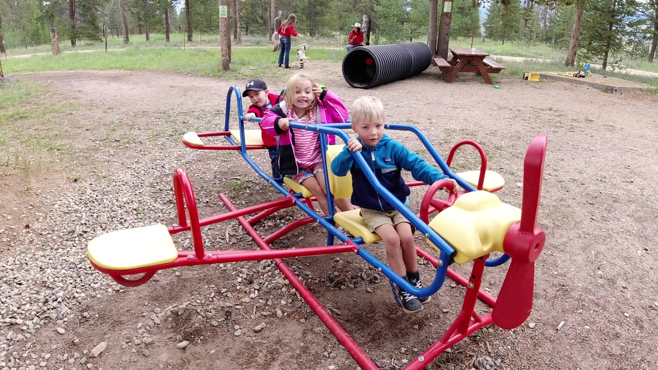 The three children remain seated on the colorful playground ride in Winter Park, Colorado, with the boy at front near the red propeller and the wooded park behind him.