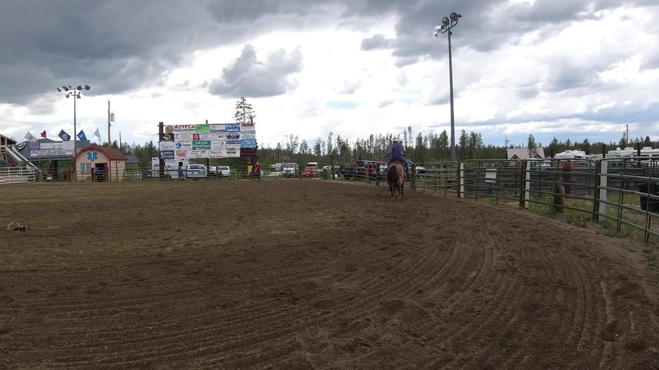Winter Park, Colorado arena view with the horse and rider slightly farther across the dirt, rail fencing on the right, bleachers, sponsor signage, parked vehicles, trees, and gray clouds.