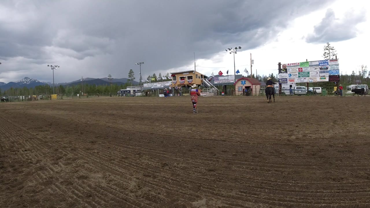 Wide dirt arena with riders, bull, buildings, and storm clouds.