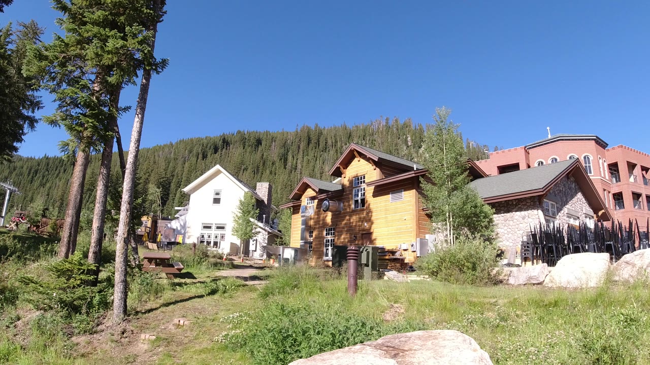 Wide hillside view with houses, trees, grass, and rocks under a blue sky.