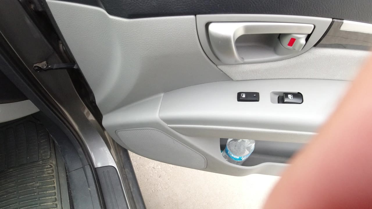 Open car door interior with gray panel, handle, switches, floor mat, and water bottle.