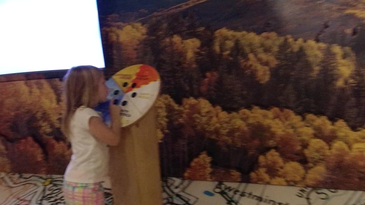 Young girl facing an interactive display wheel beside a museum wall screen.
