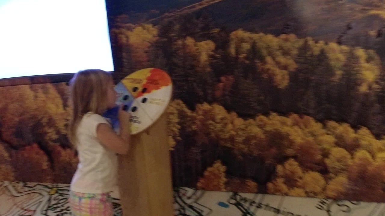 The girl reaches toward the colored museum display wheel as the bright monitor hangs above the forest mural in the Wheat Ridge exhibit area.