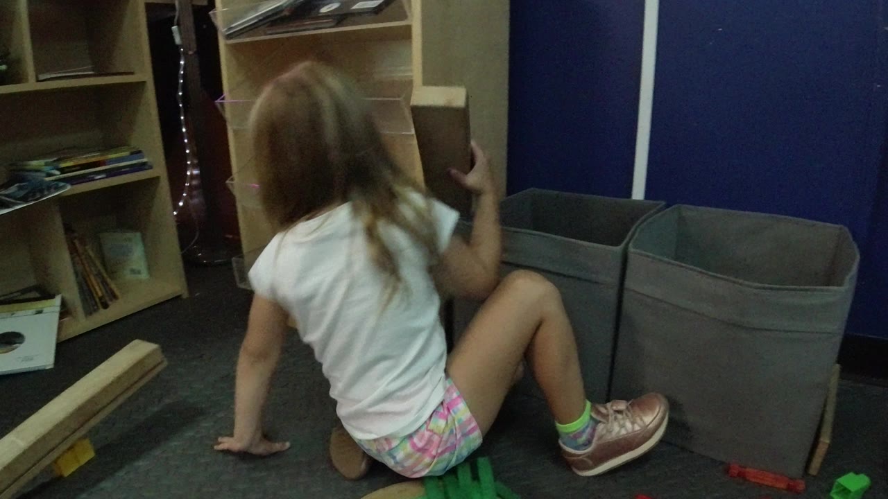 The child remains seated on the carpet in Wheat Ridge, Colorado, turned partly away from camera with shelves, papers, gray bins, and a long wood piece around her.