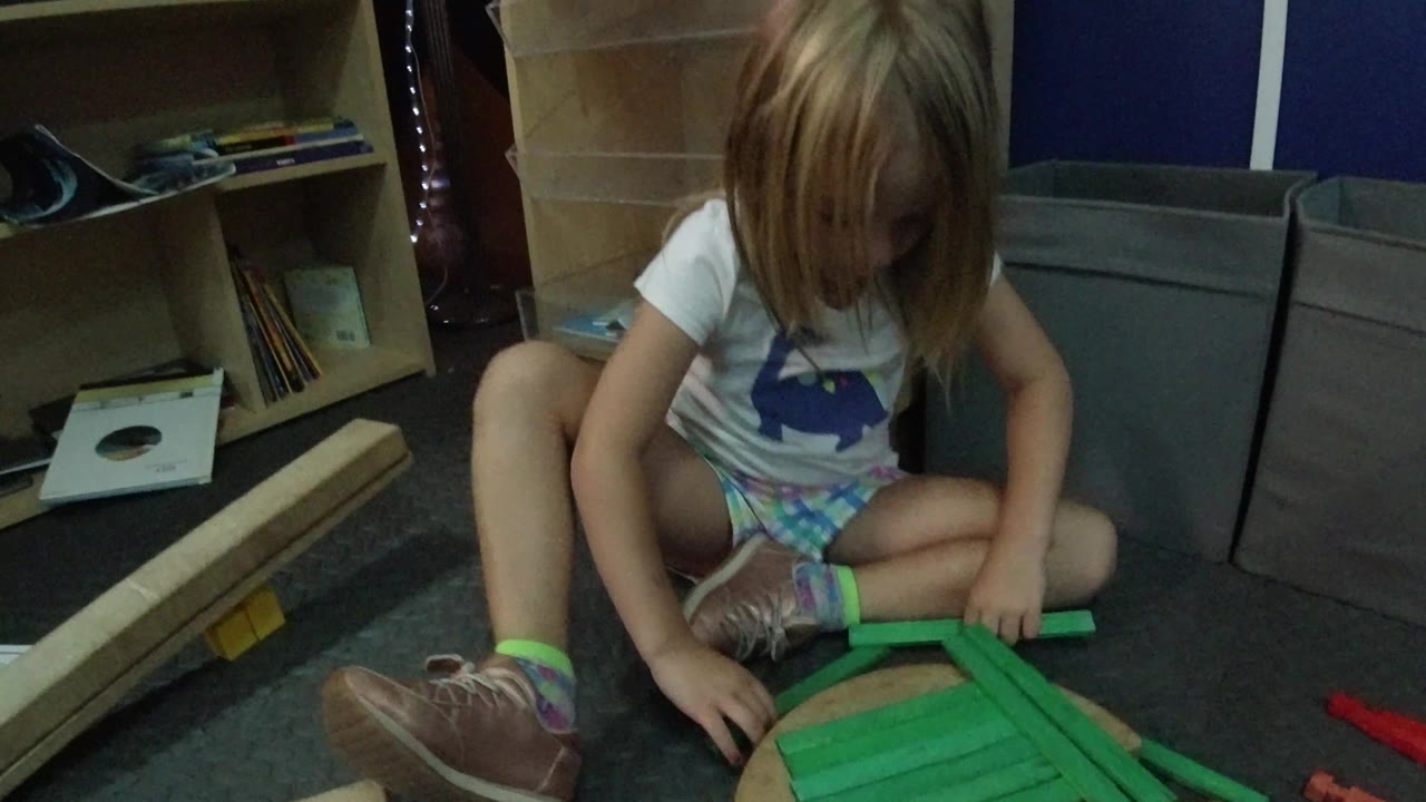In Wheat Ridge, Colorado, the girl leans over the green block structure with one shoe close to the pieces and shelves of books and toys behind her.