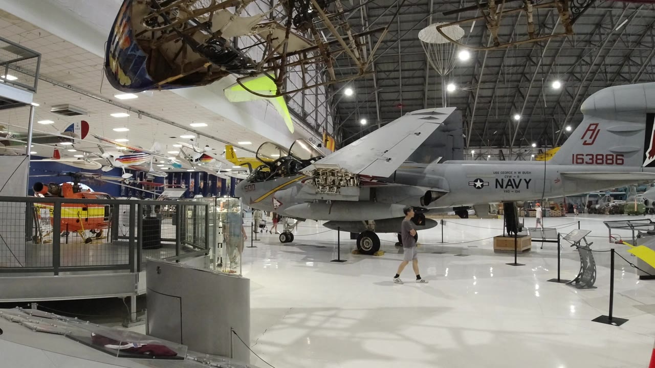 Wide indoor aerospace exhibit with a gray Navy aircraft and visitors.