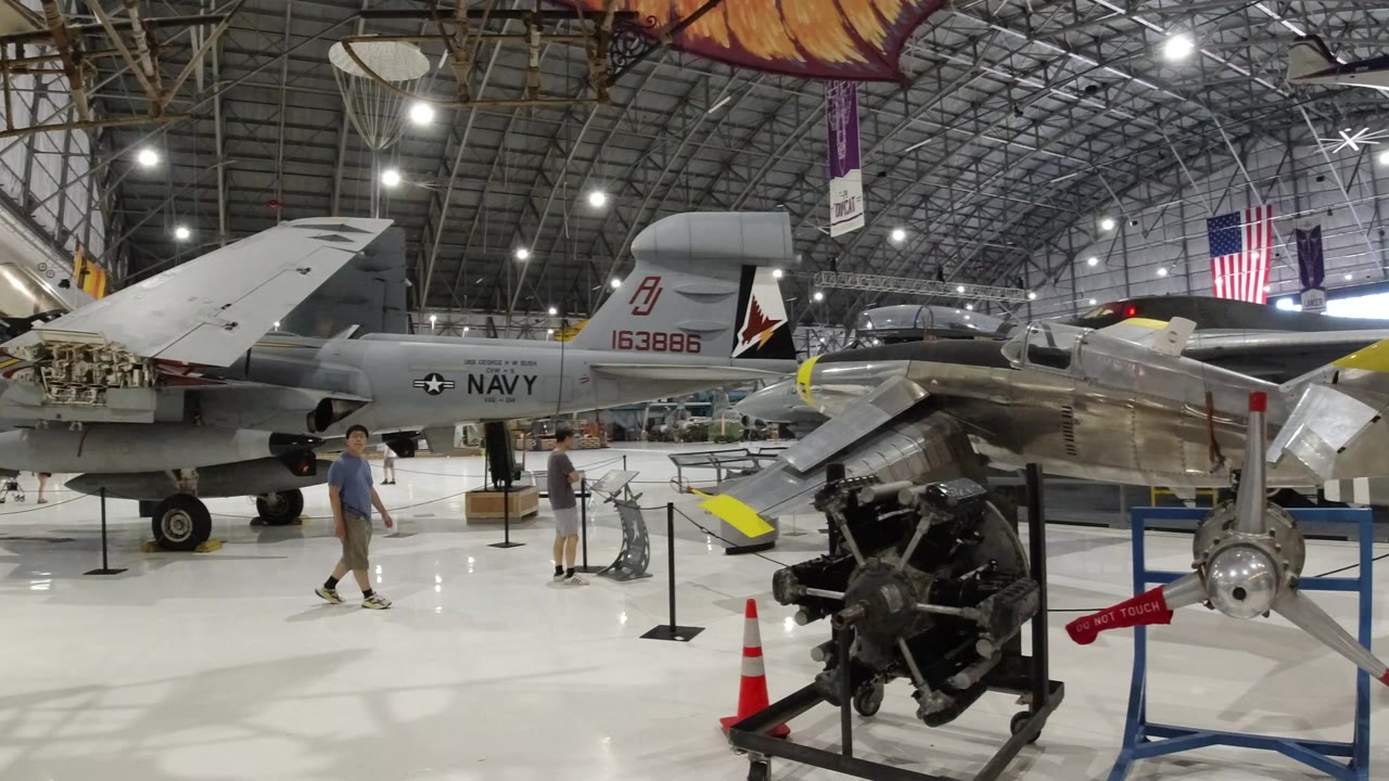 Inside a Wheat Ridge, Colorado hangar, a grey Navy-marked aircraft sits behind ropes while an exposed engine and propeller display fill the foreground with visitors visible nearby.