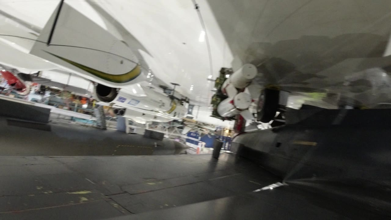 Upside-down wide view in Wheat Ridge, Colorado showing the underside of a white space shuttle display, exhibit flooring, railings, and distant visitors near the left background.