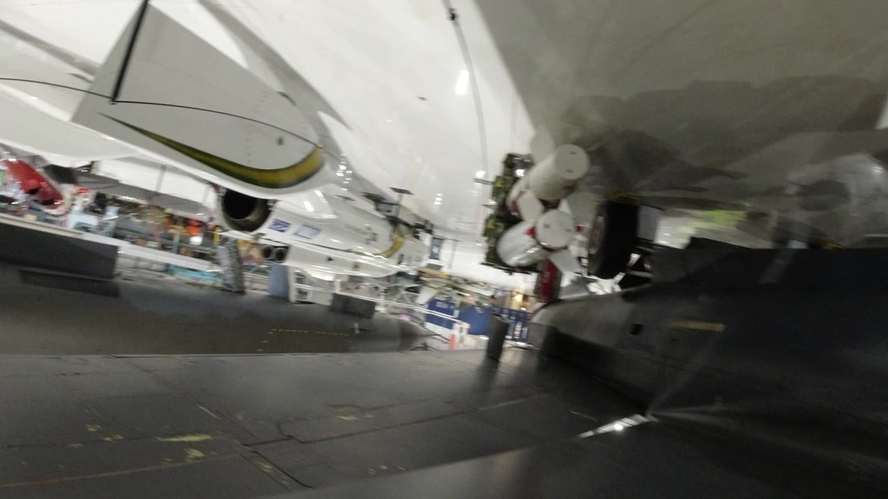 Upside-down Wheat Ridge museum frame with the shuttle underside filling the upper view, cylindrical booster components at right, and exhibit barriers and visitors deeper in the hall.