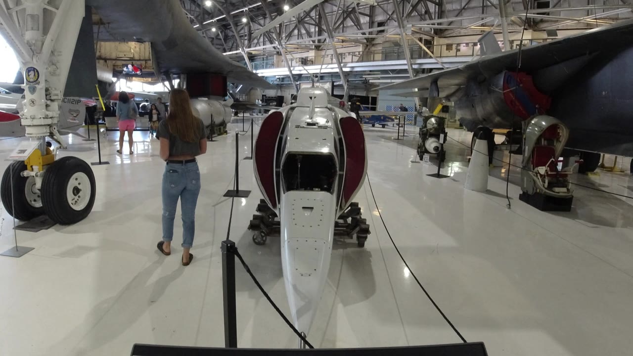 Wheat Ridge, Colorado aircraft exhibit with the white pod centered, landing gear at left, a dark aircraft body overhead, and distant visitors in the hangar.