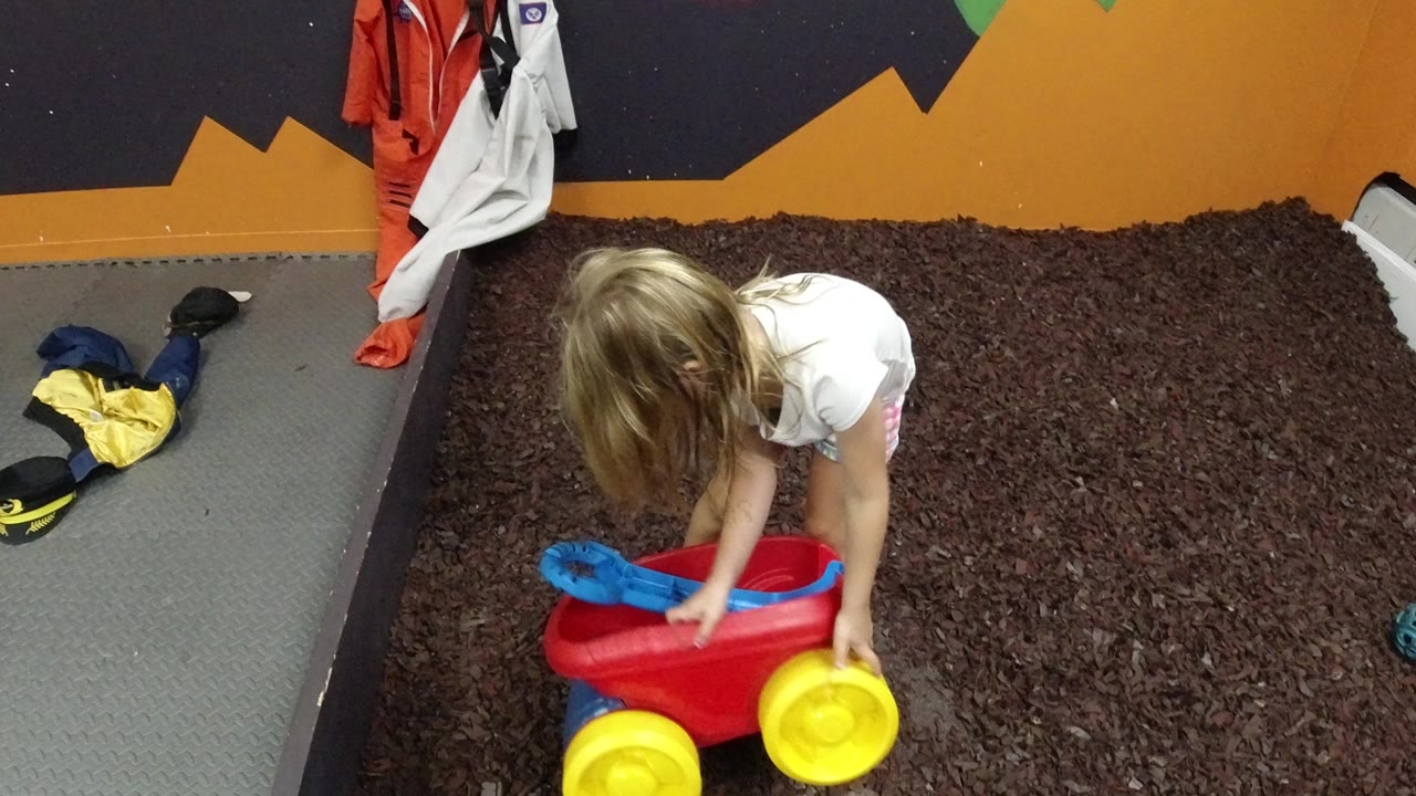 Young girl bending over a plastic wagon on brown carpet.