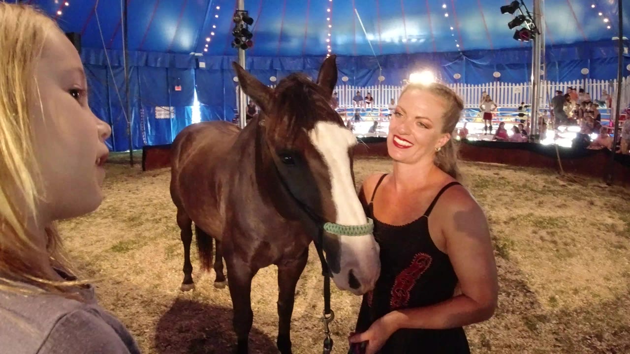 Young girl beside a brown and white horse and smiling woman inside a circus tent.