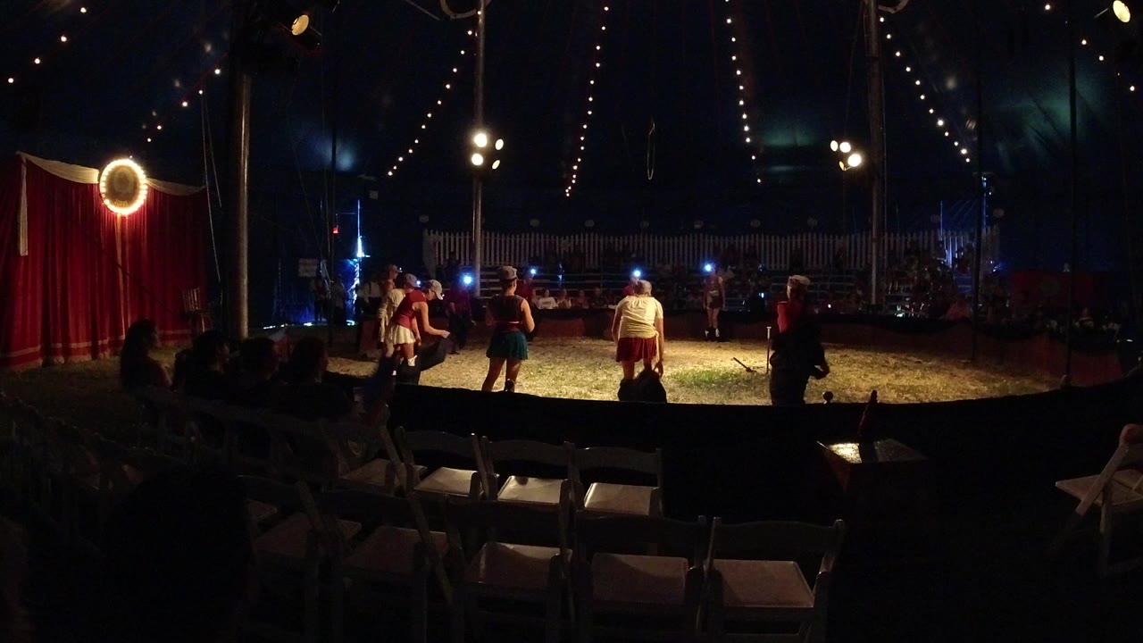 Wide view inside a Broomfield, Colorado circus tent with empty folding chairs in the foreground, red curtains at left, and several costumed performers standing in the lit ring.