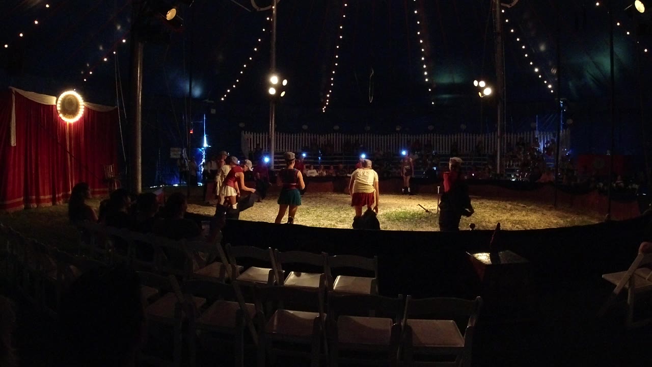 Several performers in circus costumes gather under warm lights in the Broomfield tent while the empty audience chairs remain dark in the foreground.