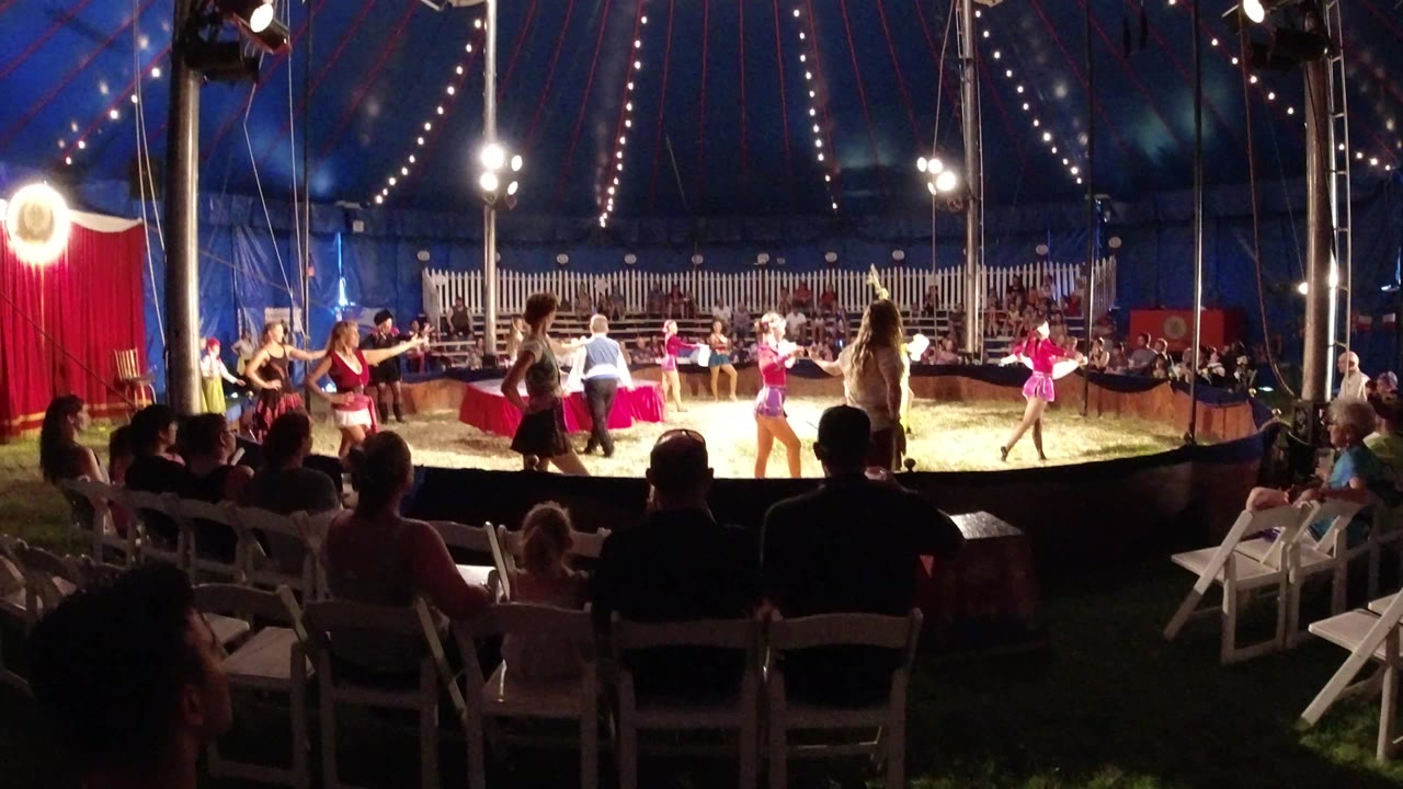 Audience members sit in the foreground while costumed performers in pink and red move around the circular performance area under bright tent lights in Broomfield, Colorado.