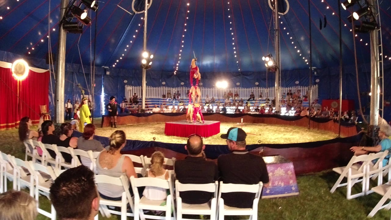 Audience members in Broomfield, Colorado sit on white folding chairs and clap while acrobats balance on a red platform under bright circus tent lights.